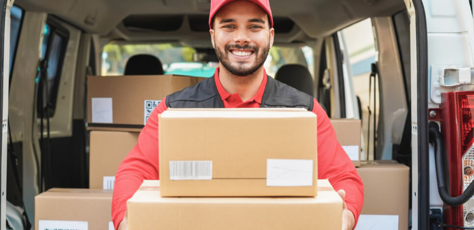 Entregador de encomendas segurando uma pilha de caixas enquanto sorri com uma van de entrega ao fundo
