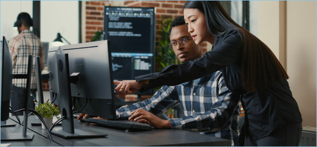 Homem negro e mulher asiática sentados em frente ao computador enquanto a mulher aponta para os códigos na tela