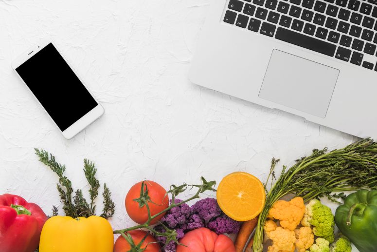Um notebook, em celular e frutas e legumes sobre uma mesa branca.