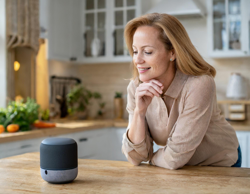 a imagem mostra uma mulher de meia idade na cozinha da sua casa. Ela está comprando itens para sua casa através de um comando de voz.