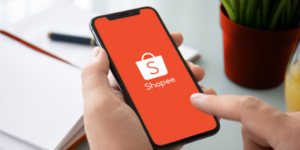 a imagem mostra uma mão segurando um celular conectado à tela inicial do App da Shopee