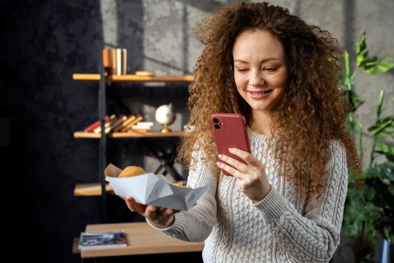 Mulher sorridente segurando lanche e tirando foto com celular, representando estratégia de clickbait visual no e-commerce.