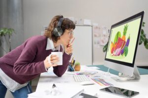 Mulher usando computador para criar design gráfico no Canva, com expressão concentrada e materiais gráficos na mesa
