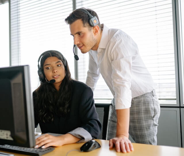 a imagem mostra uma mulher sentada com um headset na frente de um computador. Ao lado dela está um homem também com um headset e os dois estão conversando.