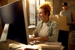 A imagem mostra uma mulher sentada em frente de um computador. Ela está sorridente pois está vendendo mais.