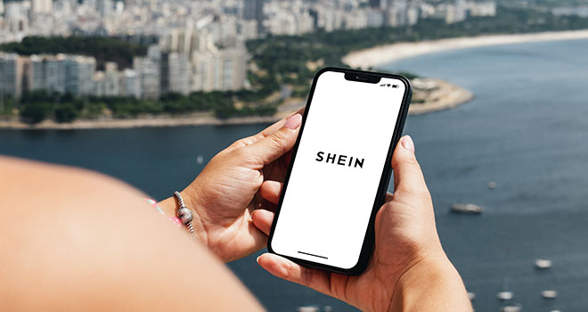 Mão feminina segurando um smartphone com a logo da shein com uma vista de uma praia ao fundo