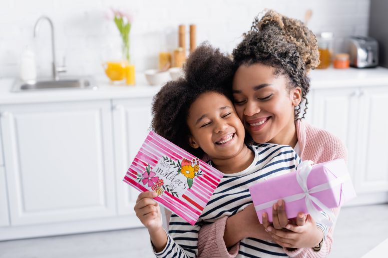 Mãe e filha sorrindo com cartão e presente em mãos — imagem representando ideia de presente para quem busca o que vender no Dia das Mães.