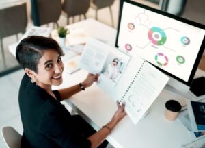 Mulher analisando relatórios gráficos impressos e digitais em ambiente de trabalho, representando o uso da visão sistêmica nos negócios.