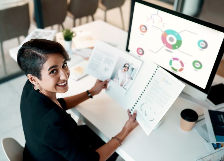 Mulher analisando relatórios gráficos impressos e digitais em ambiente de trabalho, representando o uso da visão sistêmica nos negócios.