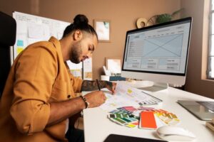 Profissional de marketing criando layouts de campanha, com amostras de cores e wireframe de site exibido no computador.