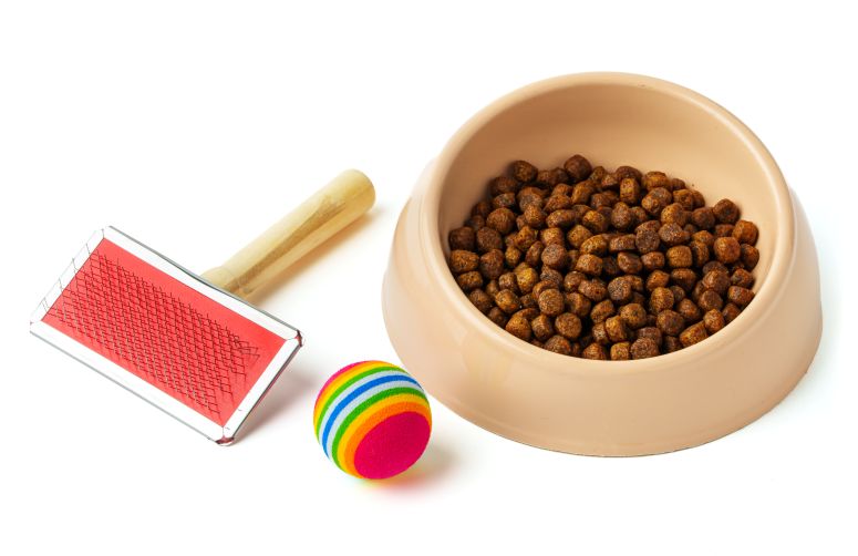 Comedouro com ração, escova para pelos e bola colorida para pets, representando itens para kits de produtos pet.