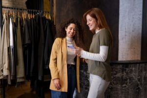 Duas mulheres jovens, estão sorridentes dentro de uma loja olhando a para a tela do celular