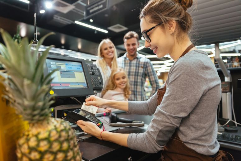 A imagem mostra uma família fazendo comoras em um supermercado e atendente passado as compras sorridente