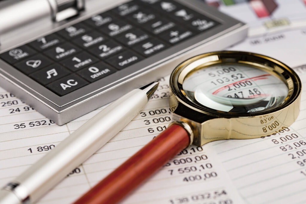 "A imagem mostra uma lupa, uma caneta e uma calculadora sobre um documento com muitos números. A lupa está em destaque, ampliando alguns valores para análise.