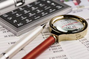 "A imagem mostra uma lupa, uma caneta e uma calculadora sobre um documento com muitos números. A lupa está em destaque, ampliando alguns valores para análise.