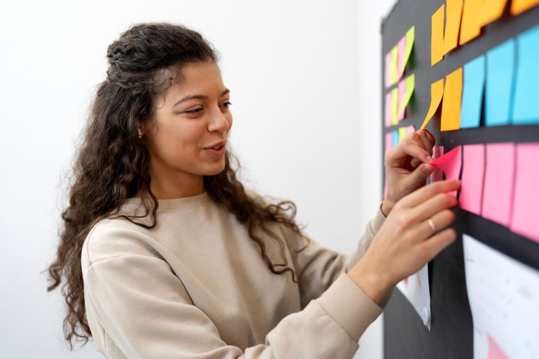 Mulher organizando tarefas com post-its coloridos em quadro de planejamento, simbolizando a criação de um fluxo de trabalho eficiente.