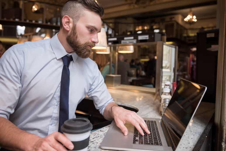 Pessoa digitando em um notebook sobre uma mesa verde ao ar livre, com um copo de café ao lado, em um ambiente iluminado pelo sol.