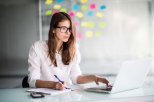 Jovem empresária anota atentamente as sua dúvidas enquanto olha para tela do seu notebook