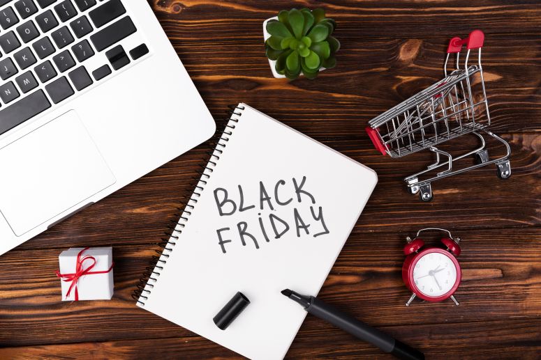 Um bloco de notas escrito Black Friday, um notebook aberto, e um carrinho de compras em mini atura.